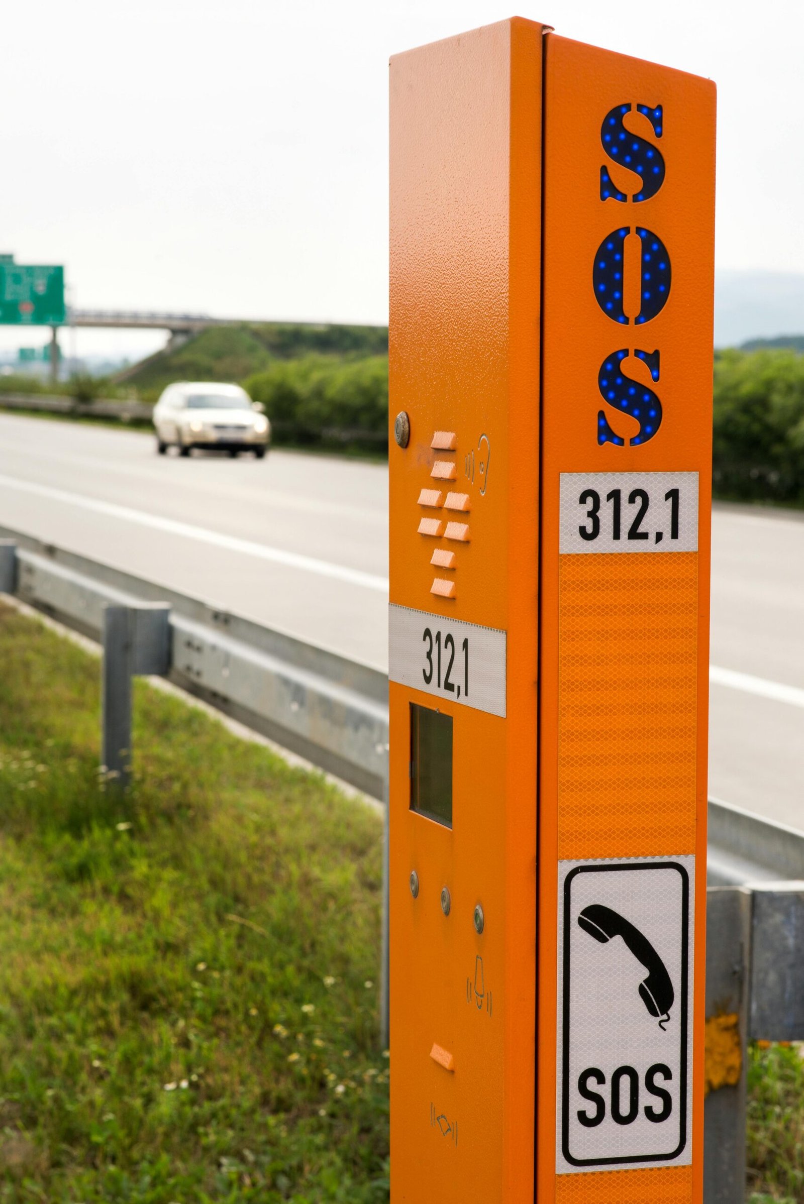 Society SOS Emergency App Orange SOS emergency phone on a highway with a blurred car in the background.