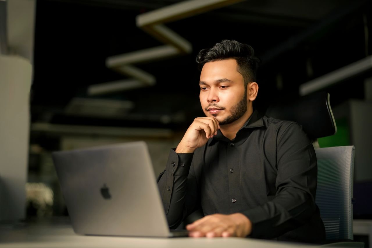 Young South Asian man in black shirt concentrating on work with laptop in modern office.
